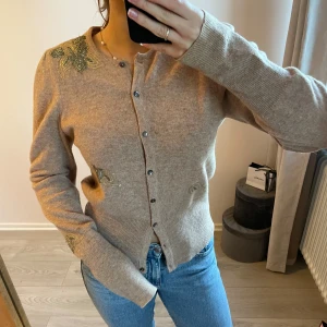 Beige kofta med pärlbroderi - Säljer en beige kofta med rund hals och knappar framtill. Från märket French connection. Koftan har dekorativa pärlbroderier i form av blad på axel, ärm och sida. Materialet är 85% ull. Koftan har lite små defekter såsom lite missfärgning på bild 4, en knapp som saknas och lite små hål som knappt syns. 
