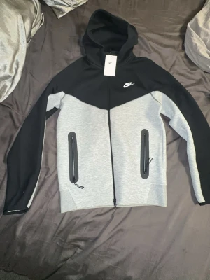 Nike Tech Fleece hoodie svart/grå - Nike Tech Fleece hoodie i svart och ljusgrått med dragkedja framtill och två stora fickor med svarta dragkedjor. Klassisk Nike-logga på bröstet och huva för extra stilpoäng. Perfekt för en sportig och avslappnad look.