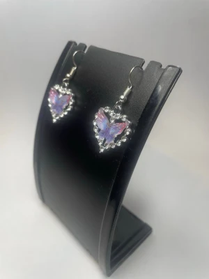 Hjärthängande örhängen med fjäril - Snygga örhängen med hjärtformad design och en lila-rosa fjäril i mitten. Runt hjärtat sitter små klara pärlor som ger extra bling. Krokar i silverfärgad metall. Perfekta för dig som gillar söta och unika accessoarer.