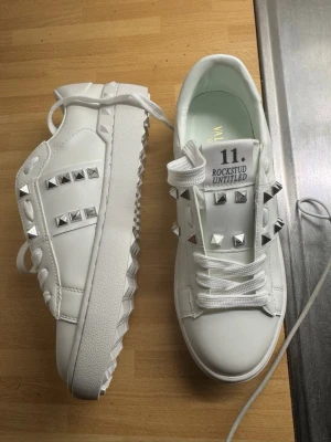 Valentino Rockstud vita sneakers - Snygga vita sneakers från Valentino med silverfärgade nitar och coola detaljer. Modellen heter Rockstud Untitled och har en chunky sula samt logga på plösen. Skorna är i läder och har klassisk snörning. Perfekta för dig som vill sticka ut med stilrena men edgy skor.