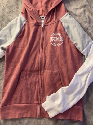 Rosa och grå hoodie från PINK - Säljer en snygg hoodie från PINK med dragkedja framtill. Tröjan är rosa med ljusgrå ärmar och har en stor ficka på magen. Trycket 'LOVE PINK' syns på bröstet. Hoodien har huva och är tillverkad i mjukt bomullsmaterial.