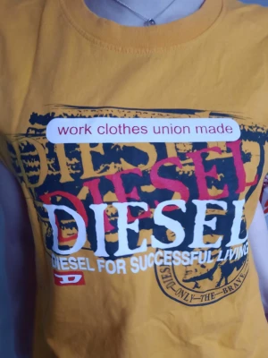 Gul Diesel t-shirt med tryck - Cool gul t-shirt från Diesel med stort tryck framtill i svart, vitt och rött. Texten 'work clothes union made' och 'Diesel for successful living' ger en streetwear vibe. Rund halsringning och ärmlös design i mjuk bomull. Perfekt för dig som gillar statement-plagg.