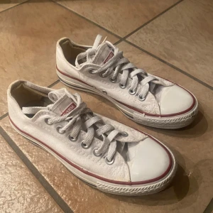 Vita Converse All Star sneakers - Klassiska vita Converse All Star low-top sneakers med röd och blå rand runt sulan. Skorna har vit canvas, vita skosnören och metallfärgade öljetter. Ikonisk logga på plösen och hälen. Perfekta för en avslappnad och tidlös stil.