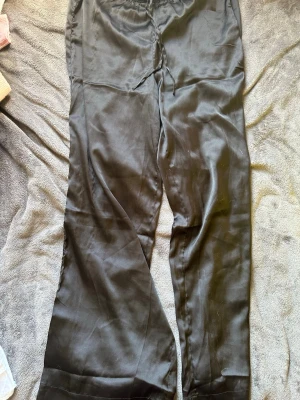 Svarta mjukisbyxor i satinlook - Säljer ett par svarta mjukisbyxor med glansig satinlook. Byxorna har resår i midjan och dragsko för skön passform. Vida ben och avslappnad stil som funkar till sneakers eller slides.