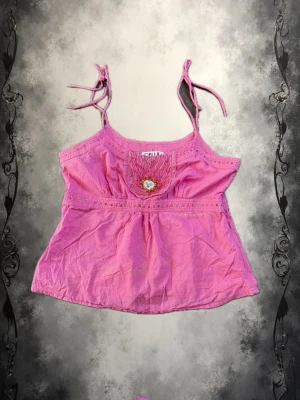 Rosa linnetopp från Joelle med pärlor - Söt rosa linnetopp från Joelle med tunna axelband som knyts upptill. Toppen har broderade detaljer och pärlor framtill samt ett färgglatt hjärtmotiv. Den har även en dragkedja i sidan ! Perfekt för dig som gillar färg och unika detaljer.