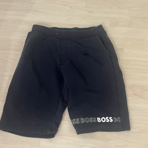 Svarta shorts från BOSS med tryck - Säljer ett par svarta shorts från BOSS med vit logotyptryck längs nederkanten. Shortsen har elastisk midja och två sidofickor. Perfekta för en avslappnad och sportig stil.