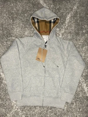 Grå hoodie från Burberry med dragkedja - Snygg grå hoodie från Burberry med klassiskt rutigt foder i huvan och dragkedja framtill. Broderad logga på bröstet och två fickor. Perfekt för dig som vill ha en stilren och lyxig look. Materialet är mjuk bomullsblandning.