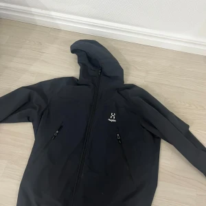 Svart vindjacka från Haglöfs med huva - Snygg svart vindjacka från Haglöfs med hel dragkedja framtill och två praktiska fickor med dragkedja. Jackan har en justerbar huva och minimalistisk design med Haglöfs-logga på bröstet. Perfekt för blåsiga dagar och outdoor-aktiviteter.