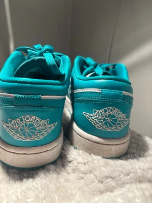 Turkosa Air Jordan 1 Low sneakers - Säljer ett par Air Jordan 1 Low sneakers i en fräsch turkos och vit färgkombination. Skorna har klassisk broderad Air Jordan-logga på hälen och Nike swoosh på sidan. Tillverkade i läder med matchande turkosa skosnören och vit sula. Perfekta för dig som vill sticka ut.