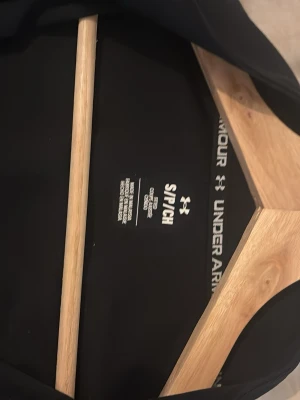 Svart träningsjacka från Under Armour - Snygg svart träningsjacka från Under Armour med hel dragkedja framtill och vit detalj över bröstet. Jackan har ståkrage och logga på vänster sida. Perfekt för träning eller uppvärmning. Tillverkad i ett lätt och funktionellt material. Byxor ingår 