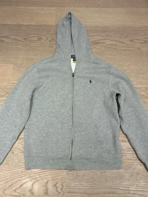 Grå hoodie från Polo Ralph Lauren - Snygg grå hoodie från Polo Ralph Lauren med dragkedja framtill och klassisk huva. Tröjan har två fickor och den ikoniska lilla loggan broderad på bröstet. Tillverkad i mjukt bomullsmaterial som är perfekt för chill dagar. Storleken är på hoodien är ”170” men funkar också som xs/s. 