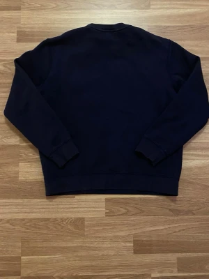 Mörkblå sweatshirt från Polo Ralph Lauren - Mörkblå sweatshirt från Polo Ralph Lauren med klassisk rund halsringning och ribbade muddar. Liten röd broderad logga på bröstet. Tillverkad i mjukt bomullsmaterial som är skönt mot huden. Perfekt för en avslappnad och stilren look.