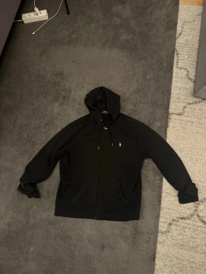 Svart hoodie från Polo Ralph Lauren - Säljer en svart hoodie från Polo Ralph Lauren med dragkedja framtill och klassisk huva med snören. Ikoniska lilla vita loggan broderad på bröstet. Hoodien har kängurufickor och är gjord i mjukt bomullsmaterial. Perfekt för en chill och stilren look.
