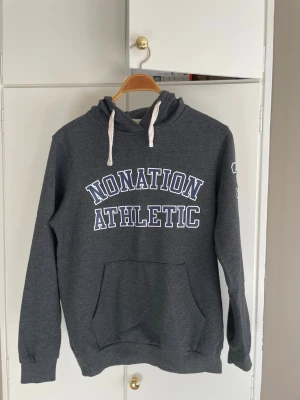 Mörkgrå college hoodie - Mörkgrå college hoodie med vit text på bröstet. Klassisk känguruficka framtill och vita dragsnören i huvan. Tillverkad i mjukt sweatshirtmaterial i storlek M. Tveka inte att höra av dig om du har några frågor.