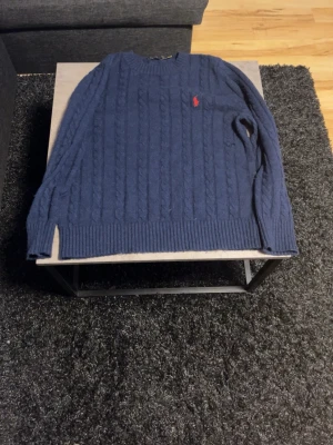 Polo Ralph Lauren stickad tröja - Kabelstickad marinblå tröja från Polo Ralph Lauren med klassisk röd logga broderad på bröstet. Tröjan har rund halsringning, ribbade muddar och nederkant samt diskreta sidoslitsar. Perfekt för dig som gillar tidlös och stilren design. OBS liten defekt vid på bild 2 men inget som syns vid användning.