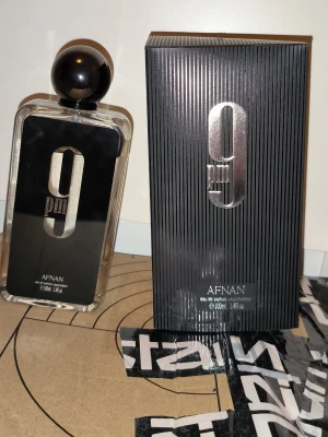 Afnan 9pm Eau de Parfum 100ml - Afnan 9pm Eau de Parfum kommer i en elegant, rektangulär glasflaska med svart och silver detaljer. Flaskan har en stor, rund svart kork och en stilren design med tydlig logga. Förpackningen är svart med silverfärgad text och räfflad yta.