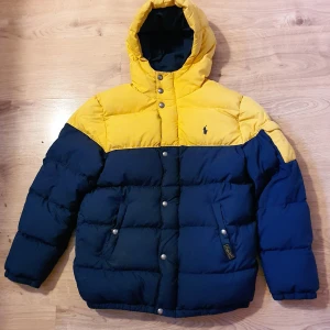 Polo Ralph Lauren blå/gul dunjacka  - Dunjacka från Polo Ralph Lauren i färgblockad gul och marinblå design. Jackan har huva, tryckknappar, dragkedja och två fickor framtill. Klassisk logga broderad på bröstet, tjock och varm jacka! Storlek 14-16 år (juniorstl L). Bra skick överlag, dock har det gula partiet blivit lite missfärgat och dragkedjan går lätt upp (man kan även knäppa med tryckknappar men på sikt är den värd en ny dragkedja).