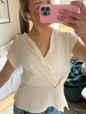Krämvit omlottopp med v-ringning - Säljer en krämvit omlottopp med v-ringning och korta ärmar. Toppen har en snygg draperad detalj framtill och lätt utsvängd nederkant. Perfekt att styla med jeans eller kjol för en chic look. Liten i storleken. Använd enbart en gång.