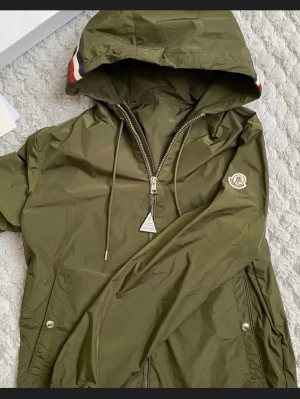Olivgrön vindjacka från Moncler - Snygg olivgrön vindjacka från Moncler med huva och dragkedja framtill. Jackan har Moncler-logga på bröstet, dragsko i huvan och fickor med tryckknappar. Materialet är tunt och vattenavvisande, perfekt för blåsiga dagar den hara bara även ett jätte jätte litet hål på framsidan som inte märks av 