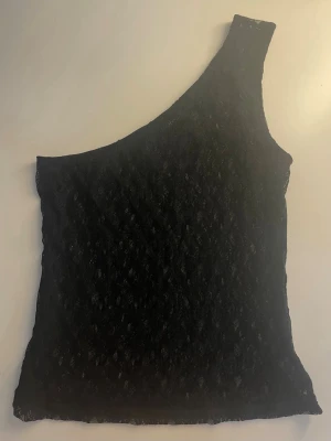 Svart spets one shoulder topp - Snygg svart topp i spets med one shoulder-design. Tunn och stretchig, perfekt för att lyfta vilken outfit som helst. Enkel modell som passar både till fest och vardag.