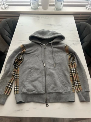 Grå Burberry hoodie med dragkedja - Snygg grå hoodie från Burberry med dragkedja framtill och klassiska rutiga detaljer längs ärmarna. Hoodien har huva med dragsko och två fickor fram. Tillverkad i mjukt bomullsmaterial som känns skönt mot huden. Perfekt för en avslappnad och trendig look.