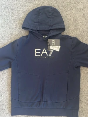 Marinblå hoodie från EA7 Emporio Armani - Snygg marinblå hoodie från EA7 Emporio Armani med stor logga på bröstet. Hoodien har huva, känguruficka och ribbade muddar. Tillverkad i mjukt bomullsmaterial som känns skönt mot huden. Perfekt för en avslappnad och trendig look. Använd endast en gång ✅ inga defekter ✅ 