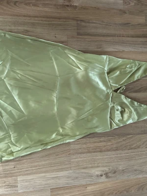 Ljusgrön kortklänning i satin - Säljer en elegant ljusgrön kortklänning i satin med smala axelband och v-ringad hals. Klänningen har en mjuk glansig yta och knyt i ryggen för extra detalj. Perfekt för sommarens alla tillfällen.