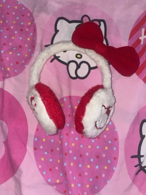 Hello Kitty öronmuffar röd/vit - Mjuka och fluffiga öronmuffar i rött och vitt med Hello Kitty-broderi på örondelarna och en stor röd rosett på bygeln. Perfekt accessoar för dig som älskar söta detaljer och vill hålla öronen varma under kalla dagar.