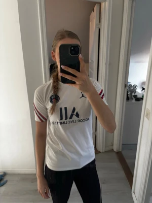 Vit PSG t-shirt  - Säljer en vit PSG t-shirt från Jordan med korta ärmar och röda samt marinblå detaljer på ärmarna. Tryckt PSG-logga på bröstet och Jordan-logga. 