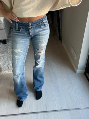Blå slitna bootcut jeans från Lee - Säljer ett par blå bootcut jeans från Lee med slitningar och hål på låren och knäna. Jeansen har klassisk femficksmodell, låg midja och ljus tvätt. Perfekta för en avslappnad och trendig look.🤍Slitna vid grenen och ett hål vid rumpan som visas på bilderna🩵