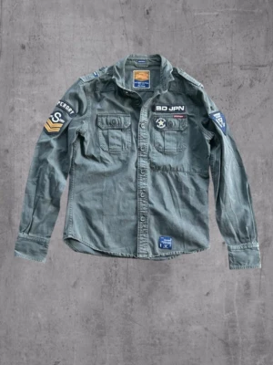 Grå overshirt från Superdry - Snygg militärinspirerad grå overshirt från Superdry med coola patches och broderade detaljer på bröstet och ärmarna. Två bröstfickor med knappstängning och klassisk krage. Perfekt för en streetwear-look.