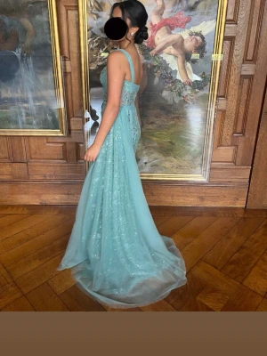 Ljusblå glittrig långklänning - Elegant ljusblå långklänning med glittriga detaljer och breda axelband. Klänningen har markerad midja och v-ringning framtill, samt ett skirt lager tyg som ger extra volym. Perfekt för bal eller festliga tillfällen. Strl 34/36. Pris kan diskuteras:)