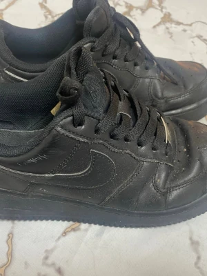 Svarta Nike Air Force 1 sneakers - Klassiska helsvarta Nike Air Force 1 sneakers i läder med svart sula och svarta skosnören. Ikonisk swoosh på sidan och perforerad tå för ventilation. Lågt skaft och tidlös streetstil som funkar till det mesta. Fina skor behöver tvättas o bli bra. Kolla bilder 