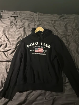 Svart hoodie från Polo Club - Snygg svart hoodie från Polo Club Saint Vincent med vit text och amerikansk flagga på bröstet. Klassisk huva med dragsko och känguruficka framtill. Perfekt för en avslappnad och sportig stil. Materialet känns mjukt och bekvämt. Den är storlek smal men finns säljer också en i medium. Skicka för mer information 