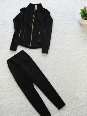 Lulemon set svart - Snyggt svart loungewear set med långärmad zip-jacka och matchande byxor. Jackan har guldfärgade dragkedjor och fickor, samt en figurnära passform. Materialet är mjukt och stretchigt, perfekt för chill dagar hemma.