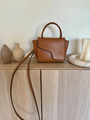 ATP Atelier väska - Snygg brun handväska i slätt läder med unik asymmetrisk lockdesign. Väskan har både ett topphandtag och en lång axelrem så du kan bära den på flera sätt. Perfekt storlek för det viktigaste och ger en trendig touch till din outfit. Små defekter. Denna färg säljs ej längre 
