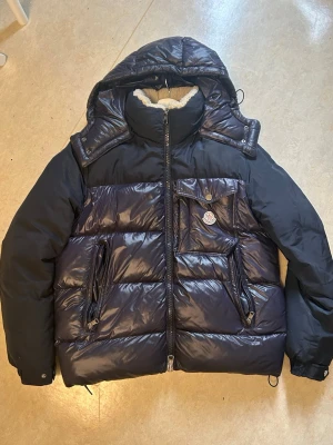 Mörkblå dunjacka från Moncler - Snygg mörkblå dunjacka från Moncler med glansig finish, avtagbar huva och mjukt teddyfoder i kragen. Jackan har två dragkedjefickor fram och en bröstficka med Moncler-logga. Perfekt för kalla vinterdagar.
