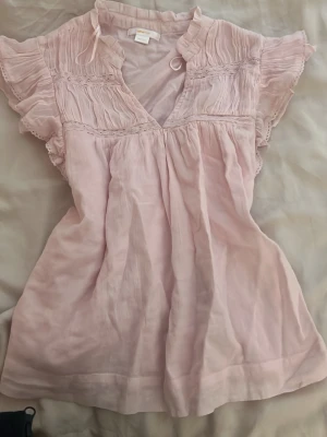 Ljusrosa blus med volangärm - Supersöt ljusrosa blus från Gina tricot med v-ringning, volangdetaljer och korta ärmar. Blusen har fina rynkade partier upptill och spetskant på ärmarna. Tillverkad i ett tunt, luftigt material som känns skönt mot huden.