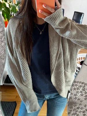 Beige ribbad kofta med öppen front - Mysig beige kofta med ribbade detaljer och öppen front. Koftan har långa ärmar och avslappnad passform, perfekt att slänga över en t-shirt. Materialet ser mjukt och stretchigt ut, vilket gör den skön att bära till jeans eller leggings.