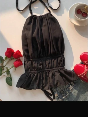 Svart volang topp med knyt - Snygg svart linnetopp med volangkant och knytband i nacken. Toppen har en rynkad midja och är ärmlös med en luftig passform. Perfekt för dig som vill ha en trendig och stilren look.