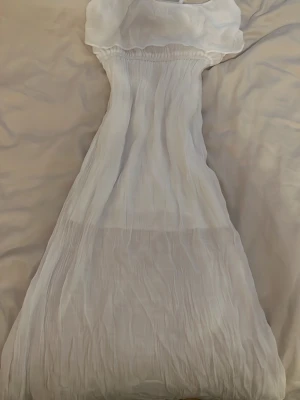 Vit långklänning med volang - Säljer en vit långklänning med axelbandslös design och volang upptill. Klänningen har en luftig och somrig känsla med resår i midjan för skön passform. Perfekt för varma dagar eller speciella tillfällen.