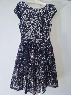 Festklänning från NELLY - Supersnygg svart kortklänning täckt med glittriga paljetter. Klänningen har korta ärmar, rund halsringning fram och en djup öppen rygg. Dragkedja bak, Perfekt för dig som vill sticka ut och glänsa på nästa fest! klänningen har en figurnära passform upptill 💥🌌