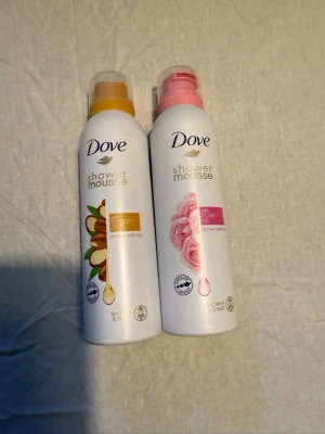 Dove shower mousse duo - Två flaskor Dove shower mousse, en med arganolja och en med rosdoft. Båda flaskorna är vita med färgglada detaljer, en med gul och brun etikett och en med rosa etikett. Perfekt för dig som vill ha en lyxig duschupplevelse.