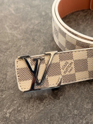 Rutig Louis Vuitton skärp i beige - Snyggt skärp från Louis Vuitton med klassiskt rutigt mönster i beige och vitt. Skärpet har en stor LV-logga i silver som spänne och är gjort i skinn. Perfekt accessoar för att lyfta din outfit.