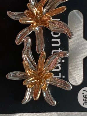 Blommiga örhängen i guld och silver - Säljer ett par snygga örhängen från Montini i form av blommor. Örhängena är i metall med både guld- och silverfärgade detaljer som ger en cool och trendig look. Perfekta för att lyfta vilken outfit som helst!
