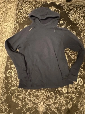 Mörkblå hoodie från Bauer - En enkel och stilren mörkblå hoodie från Bauer med huva och långa ärmar. Hoodien har en diskret logga på axeln och en magficka framtill. Perfekt för chill dagar eller när du vill ha något bekvämt på dig.