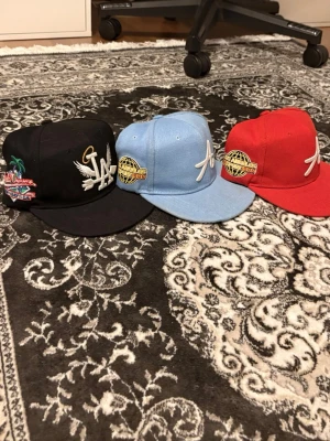 Svart LA keps med brodyr - Säljer en svart keps från New Era med LA-logga framtill, broderad vit ängelvinge och guldfärgad gloria. Sidan har färgglad brodyr med palm och texten 'World Champs'. Klassisk flat peak och justerbar baksida.