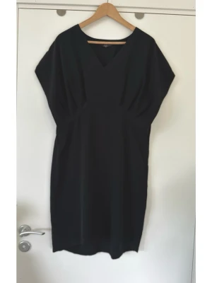 Svart v-ringad klänning från Sara Kelly, storlek 42 - Stilren svart klänning från SARA KELLY by ellos i storlek 42. Klassisk modell med V-ringning framtill och rundad halsringning baktill. Klänningen har korta ärmar och en markerad midja med resår baktill, vilket ger en smickrande passform. Designen inkluderar veckade detaljer vid bysten som bidrar till en elegant silhuett. Klänningen är knälång och har en rak nederkant. Mycket fint skick, som ny!