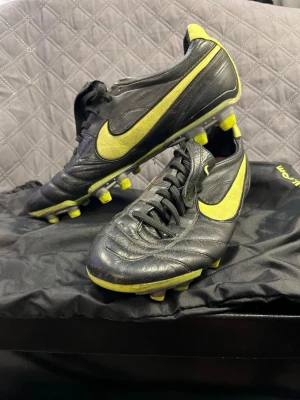 Nike Tiempo fotbollsskor - Säljer ett par klassiska Nike Tiempo fotbollsskor i svart läder med gula detaljer och gult Nike-märke på sidan. Skorna har fasta dobbar och snörning samt en vikbar plös med liten Nike-logga. Perfekta för dig som vill ha stilrena och bekväma skor på planen.