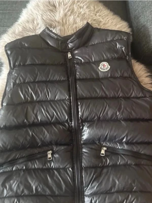 Svart dunväst från Moncler - Snygg svart dunväst från Moncler med glansig finish och klassisk logga på bröstet. Västen har ståkrage, dragkedja framtill och två fickor med dragkedja. Skick 9/10 det finns en liten skavank på baksidan men inget synligt. Skriv för intresse eller frågor!
